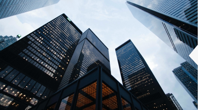 A low-angle view of tall skyscrapers with glass facades in a cityscape, set against a cloudy sky. The buildings display a grid pattern of windows, reflecting the ambient light, creating a modern urban atmosphere.