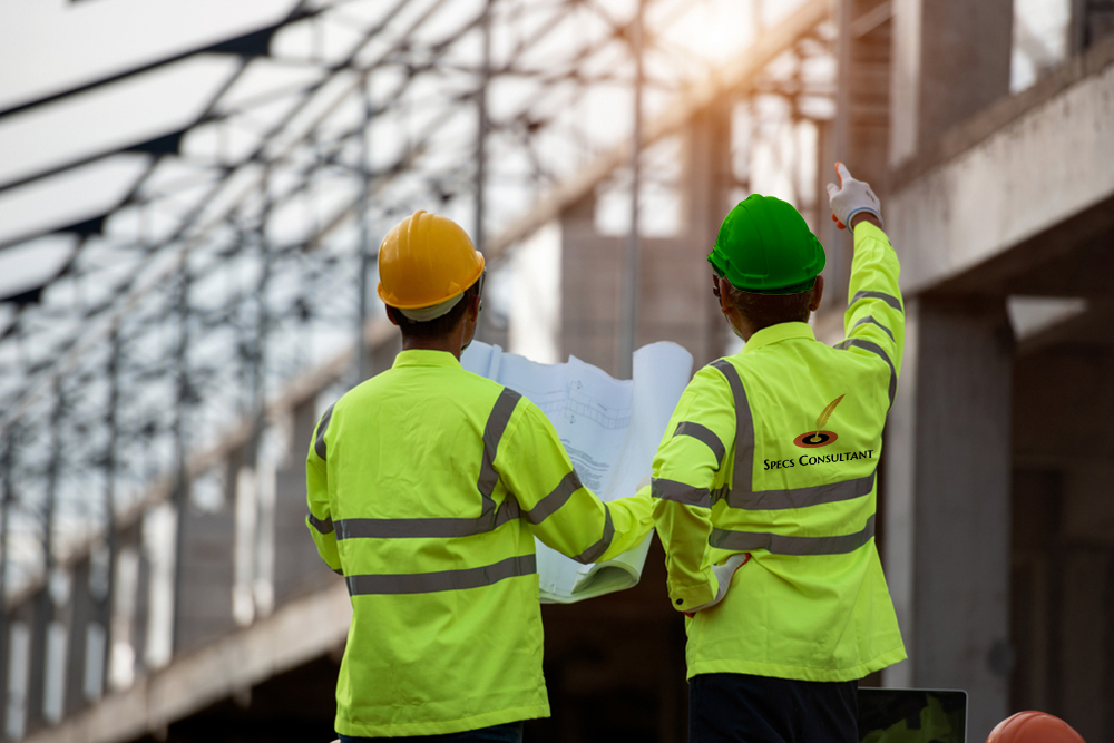 Two construction workers in yellow vests and hard hats discussing project plans on an active construction site, with building materials and equipment in the background.