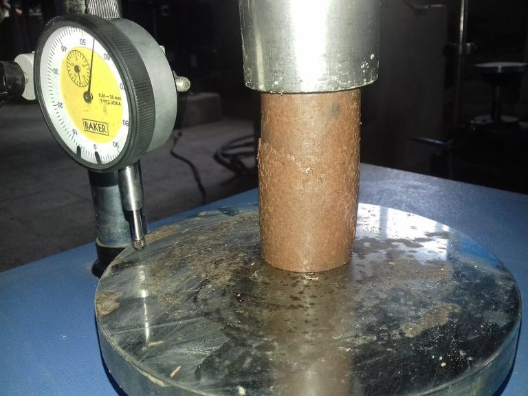 A dial indicator is positioned next to a cylindrical rusted metal pipe on a metal platform, measuring its surface. The setup appears to be part of a mechanical testing or calibration process in an industrial setting.