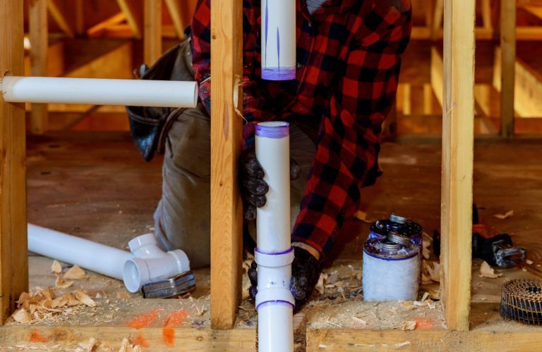 Worker wearing a red plaid shirt and gloves installs white PVC pipes within a wooden framework. Tools and plumbing materials are scattered on the floor around them.