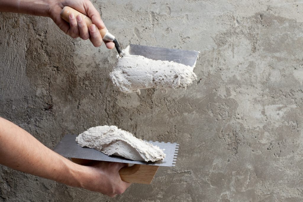 A person applies plaster with a trowel onto a rough wall. They are holding another tool with fresh plaster on it, ready to use. The wall has an unfinished texture.